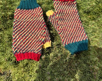 May include: A pair of knitted mittens with a colorful pattern. The mittens feature a red, yellow, and navy blue zigzag design. The cuffs and thumb are solid colors, with red, yellow, and teal accents. The mittens are displayed on a bed of green foliage.