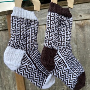 May include: Two pairs of knitted socks with a geometric pattern, hanging on wooden hangers. One pair has a white cuff, heel, and toe, with a brown and white pattern. The other pair has a brown cuff, heel, and toe, with a white and brown pattern.