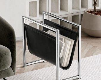 May include: A modern magazine rack with a black leather sling and a chrome frame. The rack is filled with white magazines and sits on a white rug. The background includes a gray armchair and a decorative wall.