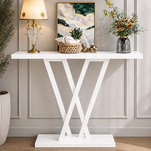 Peut inclure: Une console blanche avec une base géométrique supporte une lampe, des œuvres d'art et des objets décoratifs. La lampe a un abat-jour plissé et un pied en cristal. L'œuvre d'art présente des tons verts et dorés. Un vase de fleurs ajoute de la couleur.