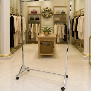 Pode incluir: Um cabide cromado com rodas está em uma boutique. O cabide está vazio, com cabides de roupas visíveis ao fundo. A loja tem uma paleta de cores neutras com detalhes dourados e um lustre.