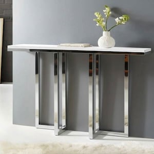 Può includere: Un moderno tavolo consolle con piano bianco e gambe cromate lucide. Un vaso bianco con fiori e un libro sono appoggiati sul tavolo. Il tavolo è contro un muro grigio.