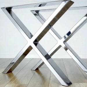 Puede incluir: Un par de patas de mesa cromadas y pulidas con un diseño en forma de X. Las patas están hechas de un metal brillante y reflectante y están diseñadas para soportar una mesa. Las patas se muestran sobre un suelo de madera clara.