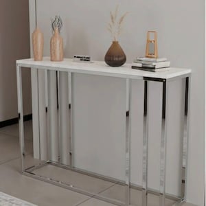 Può includere: Un moderno tavolino consolle con piano bianco e struttura in cromo lucido. Il tavolo presenta barre verticali cromate sulla parte anteriore e sui lati. Vasi decorativi e libri sono esposti sul piano. Il tavolo misura circa 122 cm di lunghezza.