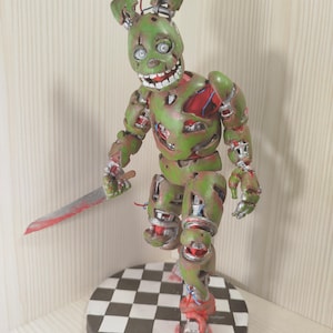 Op de afbeelding: Een groen en rood beeldje van een konijnachtig personage met een bloederig mes. De figuur heeft zichtbare mechanische onderdelen en staat op een zwart-wit geblokte basis. Het beeldje is een gedetailleerde weergave van een horrorpersonage.