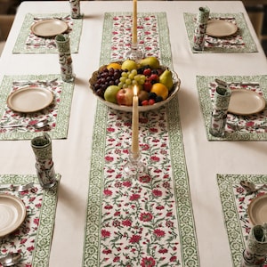 Pode incluir: Uma mesa de jantar montada para uma refeição, com uma toalha de mesa branca, um corredor floral e individuais. O corredor e os individuais têm um design floral vermelho com uma borda verde. A mesa está montada com pratos, talheres, copos e uma fruteira.