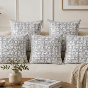 May include: Five square cushions with a grey and white floral pattern are arranged on a beige sofa. The cushions have a decorative border and are accented by a neutral-toned throw. A small vase with greenery sits on a wooden coffee table.