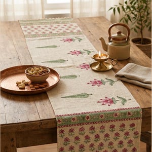 May include: A beige table runner featuring a repeating pattern of pink lotus flowers and green leaves, with decorative floral borders in green and pink. The runner is displayed on a wooden table alongside a copper tray, a bowl of nuts, a teapot, and a candle.