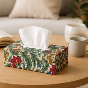 May include: A rectangular tissue box with a floral pattern in red, green, yellow, and blue. White tissues are pulled from the top opening. The box sits on a wooden table, with a mug and plant in the background.