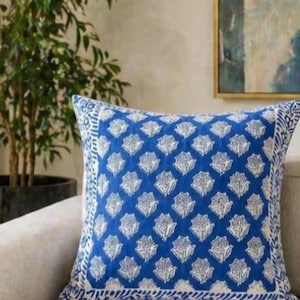 May include: A square blue decorative pillow with a white floral pattern. The pillow has a white border with a repeating design. The pillow is on a beige sofa, with a potted plant and artwork in the background.