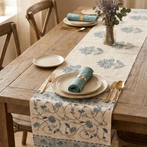 May include: A long beige table runner featuring a blue floral block print pattern and tasseled ends. The runner is laid across a wooden table set with cream-coloured plates, teal cloth napkins in gold rings, and gold-toned flatware.