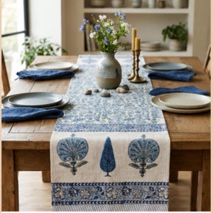 May include: A rectangular table runner featuring a cream-coloured base with blue block-printed botanical motifs, including stylised flowers and cypress trees, finished with a decorative floral border and striped edge detail on a wooden dining table.