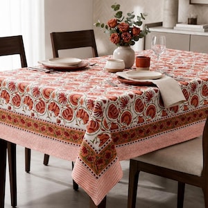 Puede incluir: Un mantel rectangular con un patrón floral repetitivo en naranja y blanco, con un borde ancho de flores naranjas y un borde fino a rayas. La tela cubre una mesa de comedor dispuesta con platos blancos, servilletas y un florero con rosas naranjas.