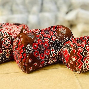May include: Three decorative, rectangular pillows with a patchwork design. The pillows feature a repeating pattern of red, white, and black geometric shapes and floral motifs on a brown background.
