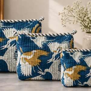 Può includere: Tre pochette trapuntate con cerniera di dimensioni decrescenti, caratterizzate da uno sfondo blu intenso con un motivo ripetuto di gru bianche e giallo senape in volo. Ogni pochette presenta un bordo in tessuto a righe bianche e nere.