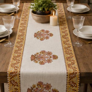 Op de afbeelding: Een beige tafelloper met een bloemenpatroon in oranje en bruine tinten. De loper ligt op een houten tafel, met couverts, glaswerk en een brandende kaars. De loper heeft een decoratieve rand.