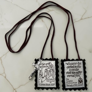 May include: A brown scapular featuring two rectangular fabric panels with scalloped black felt edges, connected by a brown cord. One panel depicts Our Lady of Mt. Carmel and St. Simon Stock with text, while the other displays the promise text.