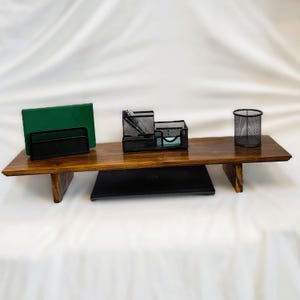 Peut inclure: Un support d'écran rectangulaire en bois avec une finition brun foncé et deux pieds de support. Sur le dessus se trouvent un carnet vert, un organisateur de bureau en maille métallique noire contenant des stylos et un pot à crayons cylindrique.