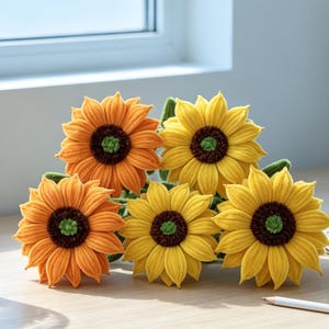 May include: A collection of five handcrafted crochet sunflowers featuring vibrant yellow and orange petals with dark brown centers and small green centers. The flowers are arranged together, showcasing detailed stitching and soft, textured fabric.