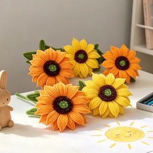 May include: A collection of five handcrafted crochet sunflowers with vibrant yellow and orange petals, dark brown centres, and green stems. The flowers are arranged on a white surface alongside a wooden rabbit figurine and a drawing of a sun.