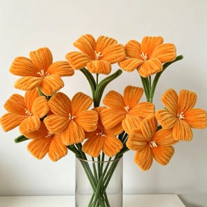 May include: A bouquet of nine orange flowers crafted from fuzzy pipe cleaners, each featuring five rounded petals and a small white center. The flowers are attached to long, thin, green stems and are arranged in a clear glass cylindrical vase.