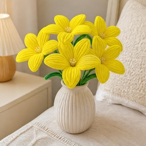 May include: A bouquet of bright yellow plush flowers with green stems arranged in a white ceramic vase. Each flower features six rounded petals with a textured surface and a small centre detail. The vase has a vertical ribbed pattern.