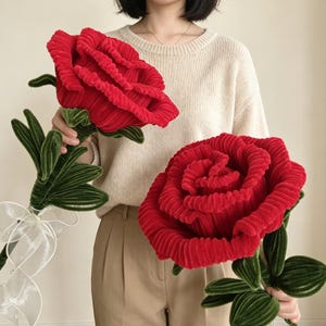 May include: Two large, plush red roses made of corduroy fabric. Each flower features layered, ruffled petals in a deep red hue, attached to a green stem with several textured green leaves. One rose is tied with a sheer white ribbon at the base.