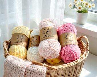 May include: A wicker basket containing four skeins of yarn in pastel yellow, white, light pink, and bright pink, each with a brown paper label reading Cotton Crochet Studio. A wooden crochet hook rests on a partially crocheted light pink fabric piece.