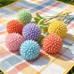 Può includere: Una collezione di sette fiori artificiali sferici in tonalità pastello verde chiaro, arancione, viola, giallo, rosa e azzurro. Ogni fiore presenta una consistenza densa e appuntita ed è fissato a un sottile stelo verde con piccole foglie.