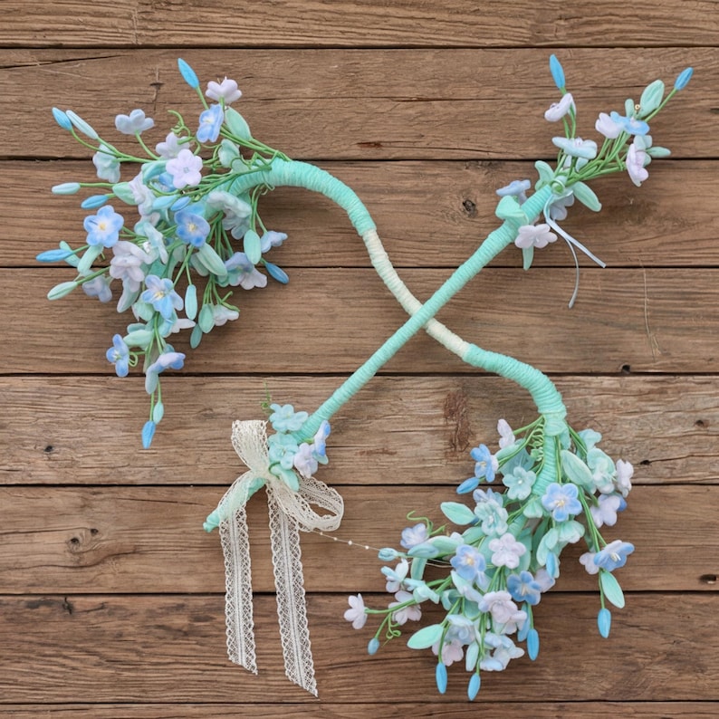 May include: Two crossed decorative wands featuring clusters of light blue and white artificial flowers. The stems are wrapped in light green material, and one wand is accented with a delicate white lace ribbon tied in a bow near the base.