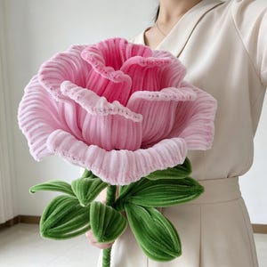 Peut inclure: Une grande rose artisanale fabriquée en tissu chenille rose côtelé et doux. La fleur présente un centre rose foncé qui se dégrade vers des pétales rose clair. Elle possède une longue tige verte avec plusieurs feuilles vertes texturées.