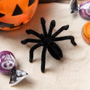 May include: A black fuzzy spider craft made from pipe cleaners with eight bent legs and a central body. The spider is positioned near an orange plastic jack-o'-lantern container and several wrapped candies with purple and orange foil wrappers.