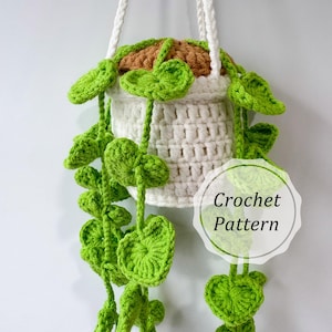 May include: A handmade crochet hanging planter with a white basket, brown soil, and vibrant green leaves. The planter is suspended by a white rope. The image includes the text "Crochet Pattern".