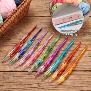 May include: A set of eight colorful crochet hooks with resin handles containing gold flakes and various colored inserts. The hooks are arranged diagonally on a wooden surface. A circular inset shows a pink case with a rose graphic and text.