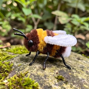 May include: A fuzzy bee figurine crafted from pipe cleaners, featuring a striped body of brown and bright yellow, white wings, black antennae, and two small black bead eyes. The insect-shaped craft is positioned with six thin black legs.