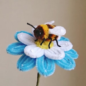 May include: A fuzzy bumblebee figurine crafted from pipe cleaners with yellow and brown stripes, black antennae, and white wings, perched atop a multi-layered flower made of white and light blue pipe cleaners with a yellow center on a green stem.