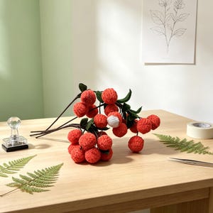 May include: A decorative branch featuring multiple crocheted lychee fruits in a vibrant orange-red hue, with dark green crocheted leaves attached to brown stems. One lychee is partially peeled, revealing a white textured interior center.
