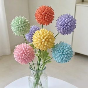 May include: A bouquet of seven spherical, spiky flowers in pastel shades of orange, light purple, yellow, light green, pink, and light blue. Each flower sits atop a thin green stem, and the entire arrangement is displayed in a clear glass vase.