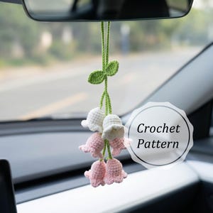 May include: A handmade crochet car charm featuring a cluster of white and pink bell-shaped flowers, suspended from a green stem with leaves. The charm is hanging from a car's rearview mirror. The text "Crochet Pattern" is visible.
