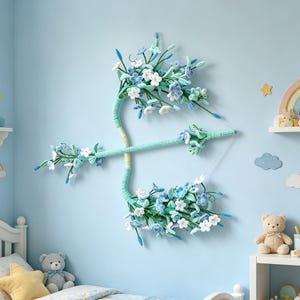 May include: A decorative wall hanging shaped like a bow and arrow, wrapped in light green yarn. The structure is adorned with numerous small, crocheted flowers in shades of white, light blue, and teal, featuring delicate green stems and leaves.