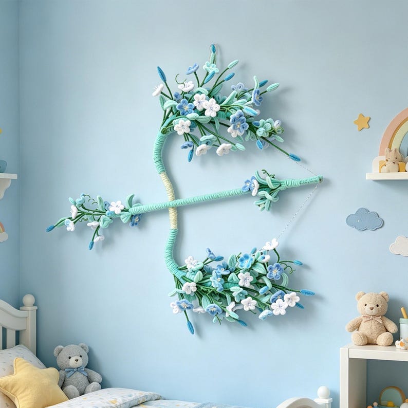 May include: A decorative wall hanging shaped like a bow and arrow, wrapped in light green yarn. The structure is adorned with numerous small, crocheted flowers in shades of white, light blue, and teal, featuring delicate green stems and leaves.