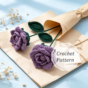 May include: Two handmade purple crochet roses with green leaves and stems are presented on a brown paper. The roses are in full bloom, and the leaves are a deep green. The text "Crochet Pattern" is visible on a tag.
