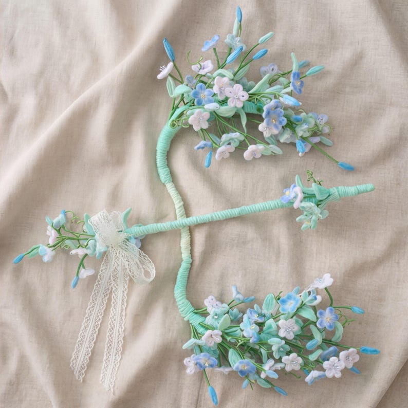 May include: A decorative hair accessory featuring two curved, mint green wrapped stems adorned with clusters of small, five-petaled light blue and white fabric flowers. A delicate white lace ribbon is tied to one side of the stem structure.