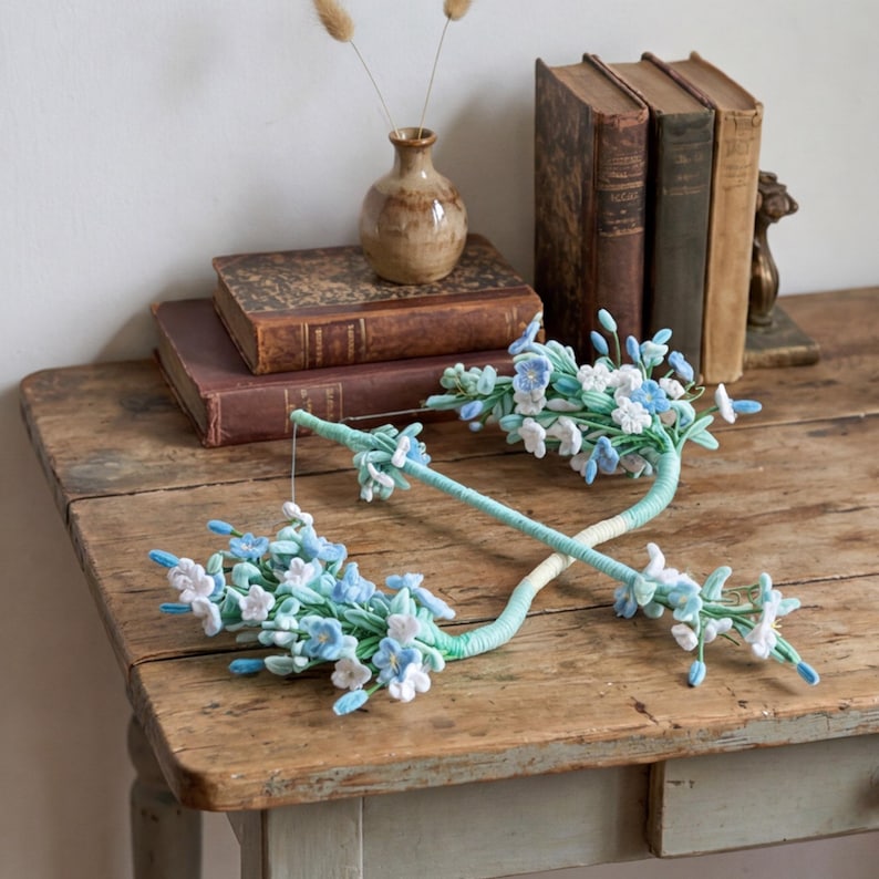 May include: Two handcrafted floral wands featuring clusters of small, light blue and white fabric flowers attached to long, mint green wrapped stems. The stems are crossed, showcasing delicate petals and buds arranged in a botanical design.
