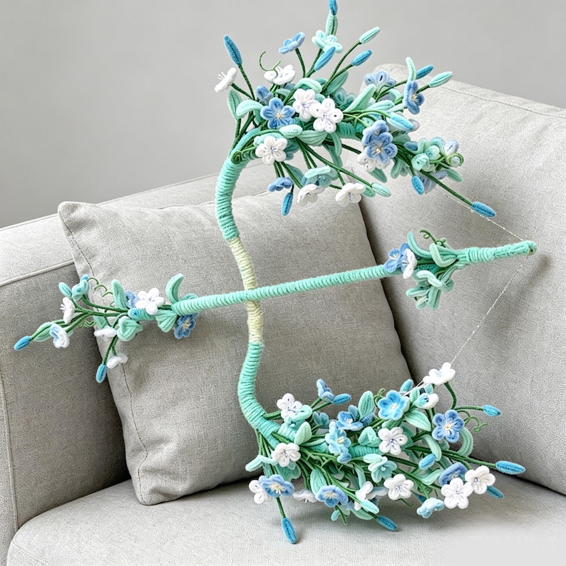 May include: A decorative bow and arrow set wrapped in light green yarn, adorned with numerous small fabric flowers in shades of white, light blue, and medium blue. The structure features green stems and leaves, with a thin string connecting the bow ends.