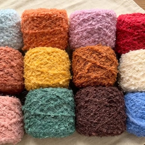 May include: A collection of twelve square-shaped yarn skeins in various colors including light blue, orange, pink, red, terracotta, yellow, brown, cream, dusty pink, teal, dark brown, and royal blue, all featuring a textured, fuzzy surface.