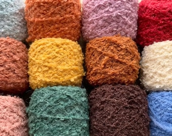 May include: A collection of twelve square-shaped yarn skeins in various colors including light blue, orange, pink, red, terracotta, yellow, brown, cream, dusty pink, teal, dark brown, and royal blue, all featuring a textured, fuzzy surface.