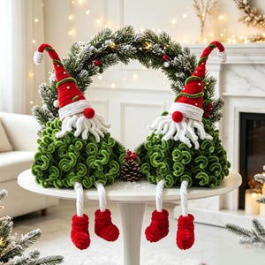 May include: Two plush gnomes with red pointed hats, white yarn beards, and green curly bodies sit on a white table. Each gnome has dangling white legs with red boots. A pine wreath with snow-dusted needles and a small pinecone sits between them.