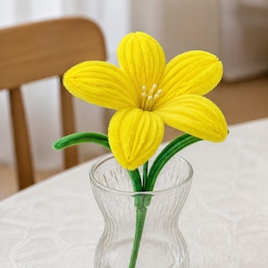May include: A bright yellow plush flower with five textured petals and a central cluster of small white stamens. The flower is attached to a green stem with two green leaves, and the entire piece is displayed inside a clear glass vase.