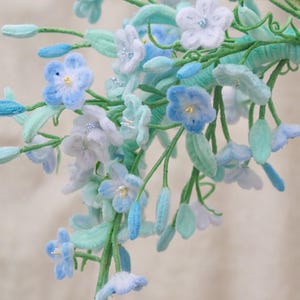 May include: A decorative floral arrangement featuring multiple five-petaled flowers in shades of light blue, white, and soft lavender. The stems and leaves are a vibrant mint green, with small, delicate buds interspersed throughout the branch.